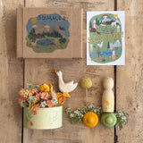 Das kleine Spieleset von Grapat und Ostheimer liegt ausgepackt auf einem dunklen Holzuntergrund. Die Verpackung - ein kleiner Karton mit einem bemalten "Hello Summer" Schuber und einer gemalten Hello Summer Karte liegen oberhalb von den einzelnen Teilen: einem bemalten Schälchen mit Blumen gefüllt, eine kleine Ostheimer Gans, eine handbemalte Figur von Grapat, 1 gelbe und eine grüne Mandala Blüte und getrocknete Blümchen liegen drapiert.