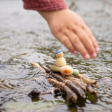 Eine Kinderhand setzt eine kleine Grapat Holzfigur von Lucky Lucky auf ein Floß in einen Bach.