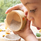 Ein Kind leert sehr konzentriert die mit Sand befüllte handbemalte Holzschale in ein kleines Tongefäß.