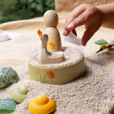 In einer Sandwanne stehen die einzelnen, handbemalten Holzfiguren von Grapat und Ostheimer, die in dem Spieleset enthalten sind. eine Kinderhand legt einen kleine Stein dazu.