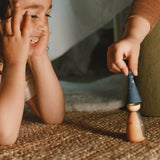 Eine Kinderhand setzt eine kegelfömige Spielfigur auf eine andere kleine Holzfigur, daneben ein anderes Kind, den Kopf in die Hände gestützt, lacht.