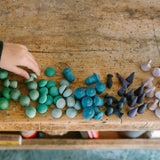 Eine Kinderhand stellt eine kleine gruene Mandalaholzfigur neben eine Vielzahl anderer Mandalasteine in blau und gruen Toenen.
