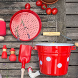 Rotes Sandspielzeug aus Metall und Emaille: ein Sieb, eine kleine, rote Schaufel mit Holzgriff, ein kleiner Kübel, rot mit weißen Tupfen. Rundherum rote und weiße Spielfiguren und rote Tomaten.