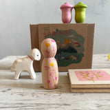 Ein Karton mit "Hello Spring" steht auf einer Holzfläche. Davor steht ein weißes Lämmchen mit einem rosa gemalten Halsband, ein länglicher rosagrün bemalter Holzkegel steht im Vordergrund. Eine Holzplatte mit Blüten bemalt liegt daneben. Auf dem Karton stehen 2 kleine, rosa und hellgrüne Holzfiguren, die wie Tulpenköpfe aussehen.