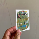 Eine Karte mit der Aufschrift Seasons! wird von einer Hand in die Kamera gehalten. Darauf ist eine geszeichnete, bunte Landschaft mit Tieren und kleinen Figuren, Wiese und Fluss, Regenbogen und Sonne.