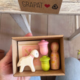 Man sieht von oben in einen kleinen Karton. Drinnen liegt ein weißes Holzlämmchen mit rosa gemalten Halsband, 2 kleine Holzfiguren in hellrosa und hellgrün, die wie Tulpenköpfe aussehen und ein bemalter länglicher Holzkegel