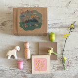 Ein Karton mit "Hello Spring" liegt auf einer Holzfläche. Davor liegen ein weißes Lämmchen mit einem rosa gemalten Halsband, ein länglicher rosagrün bemalter Holzkegel, eine Holzplatte mit Blüten bemalt und 2 kleine, rosa und hellgrüne Holzfiguren, die wie Tulpenköpfe aussehen. daneben liegen Primeln und ein Blütenzweig.