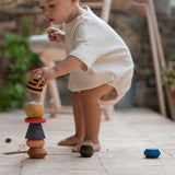 Ein Baby spielt in der Hocke mit dem Stapelturm Serentipity von Grapat. Es versucht einen gestreiften Baustein auf den Stapelturm zu stellen.