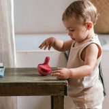 Ein Kind steht bei einem niedrigen Tisch und hält die Hand in der Luft über dem Grapat Chill Pink Bird Kreisel. Dabei beobachtet es wie der kleine Vogel-artige Kreisel sich möglicherweise bewegt hat, es sieht aus als schwankt der Kreisel am Tisch.