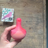 Der kleine Kreisel aus Holz Chill Pink Bird von Grapat wird in der Hand gehalten, man sieht die Vorderseite des Vogels. Am Tisch darunter liegt die Verpackung.