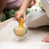Ein Kind, nur eine Hand, Zehen und ein Knie sind zu sehen, sitzt in einer Sandwanne und hält den Holzkreisel Flowing Yellow Bird von Grapat in der Hand.