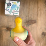 Blick von oben, eine Hand hält den Kreisel Flowing Yellow Bird von Grapat. Wir sehen die gelb, grün, bläuliche Bemalung vom Vogel-Kreisel aus Holz.Darunter am Tisch liegt die Verpackung.