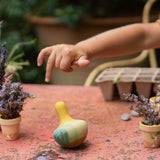 Eine Kinderhand über dem Holzkreisel Flowing Yellow Bird von Grapat, der schwankt und kreiselt. Daneben kleine Tontöpfchen mit lila Blumen.