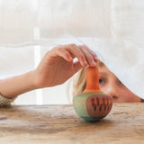 Ein Kinde hält mit seinem Mittelfinger den Mellow Orange Bird ganz vorsichtig von oben. Der Kreisel steht auf Holz, ber der Kinderhand ist eine feines Tuch, das Kind schaut unter das Tuch auf den Kreisel.