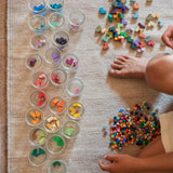 Blick von oben auf einen Teppich auf dem viele kleine Gläser stehen. Ein Kind sitzt auf dem Teppich und sortiert die bunten Tetris-artigen Steine farblich passend in die einzelnen Gläser. Lose vor dem Kind liegen nocht ganz viele Kreise, kunterbunt von Grapat Mis, Match and Bloom, daneben die eckigen Steine.