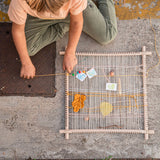 Der Grapat Frame, aufgebaut, in eine Richtungbespannt mit der Hanfschnur, liebevoll bestückt mit Blättern und Zweigen. Ein Kind webt eine gelbe Schnur ein.