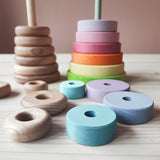 Ein Blick von vorne auf unterschiedliche Holzscheiben mit Loechern. rechts liegen die Pastellfarbenen des Grimms Scheibenturmes. Links legen die gewachsten Naturholzsscheiben vom Stapelturm Kokomoo. Im Hintergrund stehen die jeweiligen Basisplatten auf denen einige zugehoerige Lochscheiben aufgesteckt sind.