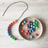In einem flachen Korb liegen Grimms Holzperlen, daneben eine Schnur auf der schon 11 Holzkugeln beginnen mit grün und blau, dann lila, pink, orange und gelb, aufgefädelt sind.