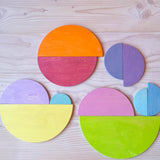 Die 11 halbkreisförmigen Bauplatten von Grimms in pastelligen Regenbogenfarben sind als Legematerial zu verschiedenen Figuren auf dem Boden aufgelegt.