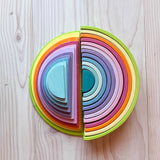 Die 11 halbkreisförmigen Bauplatten von Grimms in pastelligen Regenbogenfarben liegen übereinander gestapelt, Blick von oben, rechts daneben liegt der große Grimms Regenbogen Pastell, seitlich zu sehen.