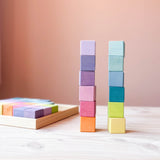 Zwei Türme mit je sechs Bausteinen vom Grimms Regenbogen Pastell Mosaik sind gebaut, links mit den Farbtönen orange, pink, rosa und lila, rechts mit gelb, grün, petrol und blau. Im Hintergrund liegt der Holzrahmen mit den restlichen Bauwürfeln.
