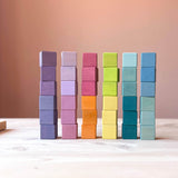 Sechs Türme mit je sechs Bausteinen vom Grimms Regenbogen Pastell Mosaik sind gebaut, ganz links mit Violett-Tönen, dann lila, der dritte Turm ist orange und pink, der vierte gelb und grün, dann petrol-Töne und ganz recht hellblau und dunkelblau.