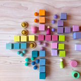 Holzbausteine vom Grimms Pastell Mosaik liegen am Bode, dazwischen kleine Filzbälle in passenden Farben, Holzbecher von Grapat und Spielfiguren aus Holz.