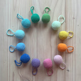 Zwölf Häkelbälle mit Schlaufen in 12 pastelligen Farben des Regenbogens, aufgelegt zu einem Kreis.