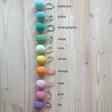 Alle zwölf Häkelbälle mit Schlaufe von Kokomoo aufgelegt in einer Reihe, daneben stehen die Farbbezeichnungen.