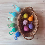 Zwölf Häkelbälle mit Schlaufen in 12 pastelligen Farben des Regenbogens, 6 Bälle in Rosa-, Lila- und Gelbtönen liegen in einem Körbchen, 6 Häkelbälle in Blau- und Grüntönen daneben.