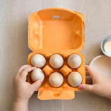 Ein offener oranger Eierkarton mit 6 naturbelassenen Holzeiern. Eine Kinderhand greift nach einem Holzei.