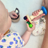 Zwei Babys spielen mit der Regenbogenrassel von Nic Toys. Ein Baby hat noch kleine Schalen in der anderen Hand.