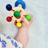 Die Regenbogenrassel Karussel von Nic Toys aus Holz wird von einer Babyhand gehalten.