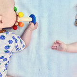 Ein Baby auf dem Bauch liegend erkundet mit dem Mund die Regenbogenrassel aus speichelecht lackiertem Holz von Nic Toys.