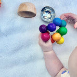 Der Kugelgreifling von Kokomoo wird von einem Baby mit seinen Händen erforscht. Daneben liegen ein kleiner Holzbecher und eine kleine Klangschale aus Edelstahl.