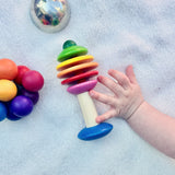 Ein kleine Klangschale, die Regenbogenrassel Baum von Nic Toys und der Kugelgreifling von Kokomoo auf einer weißen Babydecke verstreut. Eine Babyhand greift nach der Rassel.