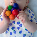 Ein Baby, auf dem Rücken liegend erkundet den bunten Kugelgreifling von Kokomoo mit Händen und Mund.