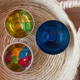 Lichtbauwürfel von Bauspiel in verschiedenen Farben liegen in kleinen Schüsseln aus Edelstahl, die in einem Korb aus Naturmaterial stehen. Ein blauer Acrylwürfel liegt in einer blauen Schüssel.