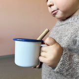 Ein weißer Becher aus Emaille mit blauem Rand. Gehalten wird die Tasse am Henkel von einem kleinen Kind.