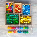 Ein Blick von oben auf 5 Pappkartons, darin befinden sich kunterbunte kleine Mandala-Holzmaterialien in verschiedennen Farben - nach Formen sortiert. Bunte Regenbogeneier, kleine orangene Kegel, Tropfen in 3 Blautönen, Kleine grüne Bäume, die wie Tannenwipfel aussehen und gelbe Honigwaben. Davor liegen von jeder Form und Farbe jeweils 1Stück aufgereiht. Daneben liegt eine kleine Mini-Holzzange, wie eine Gurkenzange, nur klein.