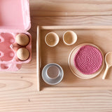 Ein Blick von oben auf ein Holztablett mit einem kleinen flachen Holzteller, auf dem ein rosa Häkeldeckchen liegt, daneben stehen 2 Holzeierbecher, liegt ein Holzkochlöffel. Neben dem Tablett steht ein rosa Eierkarton mit 2 Holzeiern.