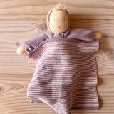 Eine Waldorfpuppe aus fliederfarbenem Nicki-Stoff liegt zugedeckt mit einem gestricktem rosa-weiß-Tuch auf einer Naturholzfläche.