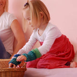 Ein Mädchen mit einem roten Tuch über ihr weißes Langarm-Shirt gebunden, kniet auf einer Matte und nimmt gestrickte Stulpen von Kokomoo aus einem Weidenkorb, der vor ihr steht.