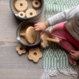 Man sieht einen Draufblick auf ein sitzendes Kind, das vor sich einen Steckturm aus Naturholz hat. Auf diesem Steckturm sind Blumenformen verschiedener Größen mit Loch auf einen Steckteil gestapelt. Einige der blumenförmigen Lochscheiben liegen neben einem Jutekorb mit grauem Rand. Daneben steht ein Korb mit runden Naturholzscheiben, mit Loch in der Mittel