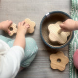 Man sieht einen Draufblick auf ein sitzendes Kind, das vor sich einen Steckturm aus Naturholz hat. Auf diesem Steckturm sind Blumenformen verschiedener Größen mit Loch auf einen Steckteil gestapelt. Einige der blumenförmigen Lochscheiben liegen neben einem Jutekorb mit grauem Rand.