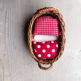 Das Wichtelnest von Kokomoo: ein ovales Weidenkörbchen als Puppenbett mit rosa getupfter Matratze, rot-weiß getupfter Decke und einem rot-rosa kariertem Puppenpolster.