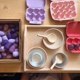 Spielset "Gruß aus der Küche" mit einem Holztablett, zwei Holzschalen, zwei Kochlöffel, einem Alugefäß, alles auf einem Tablett, dazu bunte Eierkartons.