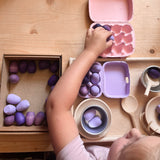 Ein Kund legt ein kleines lila Holzei in einen rosa Eierkarton. Ein kleiner lila Eierkarton ist schon befüllt. Vor dem Kind ein Tablett mit Holz Spielmaterialien.