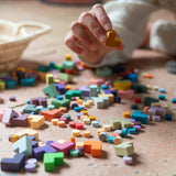 Grapat Mis Match an Bloom Steine aus Holz liegen kunterbunt am Boden. Man sieht kleine Kreise und Tetris-artige Steine aus Holz in allen Farbschattierungen. Eine Hand hält einen gelben Stein.