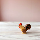 Die Holzfigur brauner Hahn von Ostheimer mit rotem Kamm steht auf einer Holzplatte. Der Hahn zum Spielen steht seitlich mit Blick nach links.